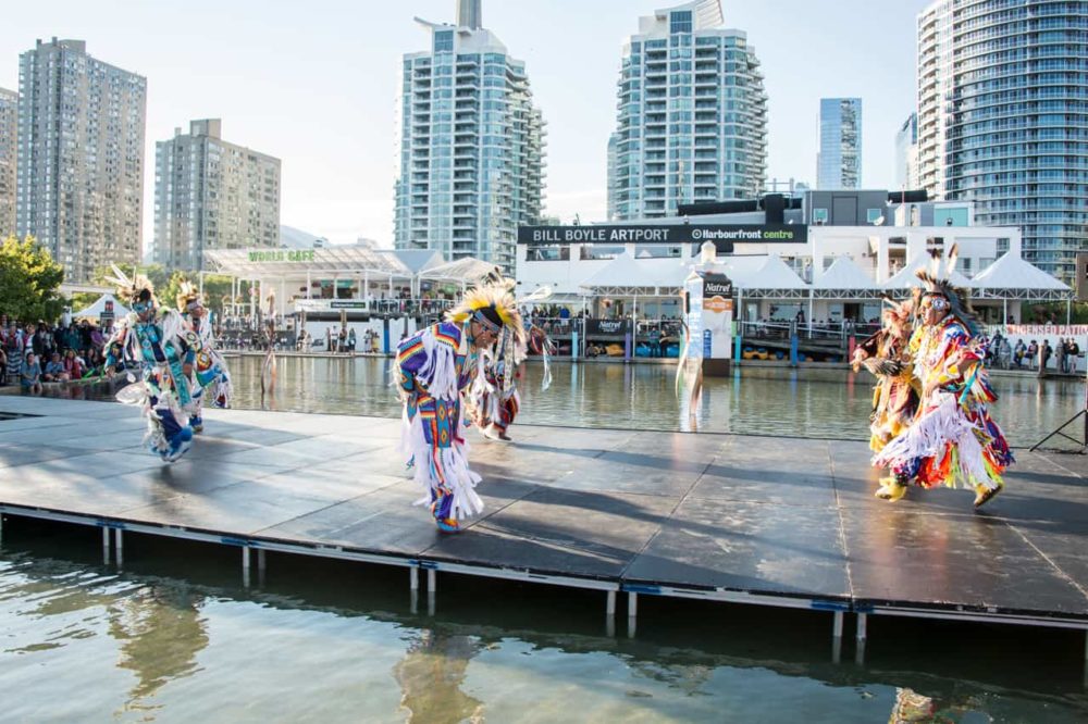 Indigenous dancers on dock overlooked by towering skyscrapers 