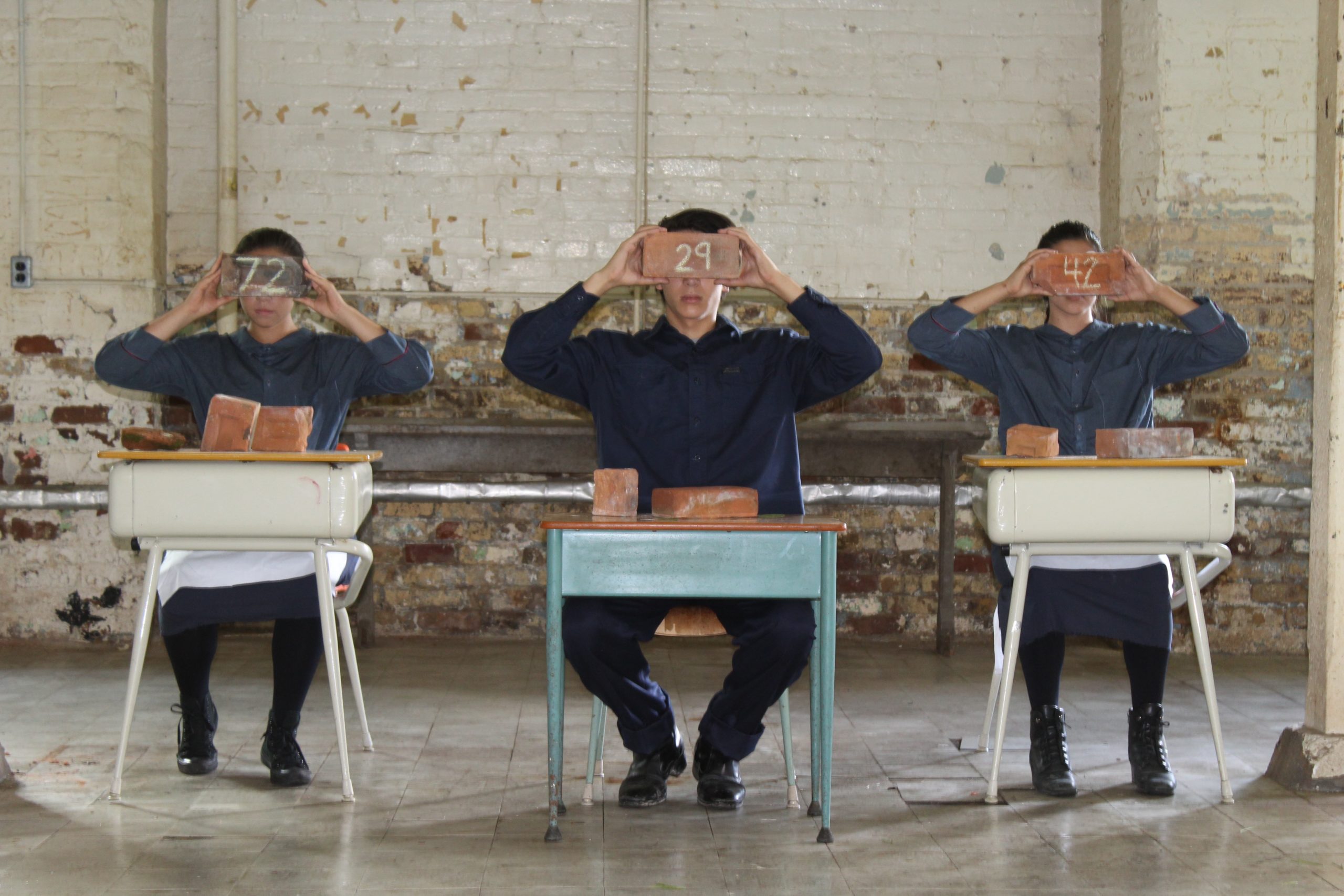 Three uniformed children sitting at desks holding numbered bricks to their forehead